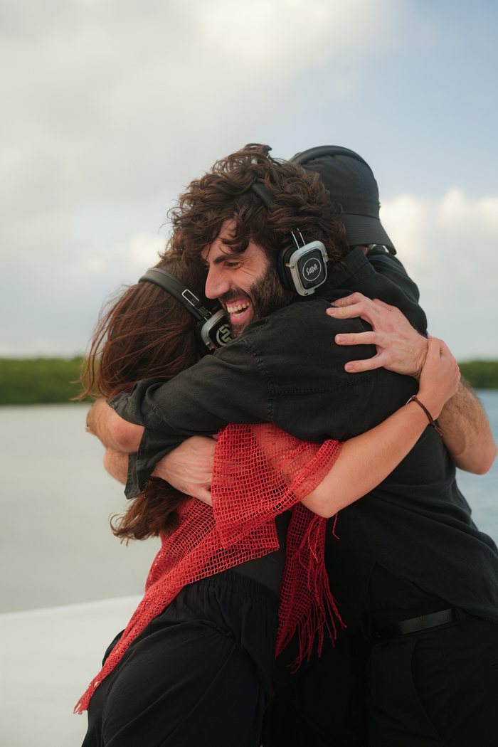 Two people in SUDA headphones holding each other in a long embrace beside the water, one wearing a red fringed scarf.